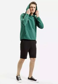 Zielona bawełniana bluza z kapturem, z kieszenią z przodu i regulowanym sznurem. Połączona z czarnymi szortami sportowymi i czarnymi trampkami.