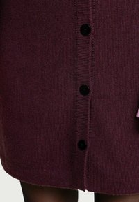 Burgunderfarbener Strickcardigan mit geradem Schnitt, der eine Reihe schwarzer Knöpfe an der Vorderseite und eine weiche Textur aufweist.