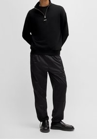 Felpa nera con zip e collo alto, con logo bianco, abbinata a pantaloni neri lucidi e scarpe nere. Design semplice e moderno.