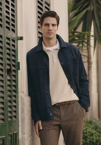 Marineblauwe suède jas met een gespreide kraag, voorknopen en borstzak, gelaagd over een beige textuurpoloshirt en bruin gestreepte broek.