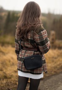 Zwarte crossbody tas met een getextureerd oppervlak, gouden logo-detail en ritsluiting. Model draagt een geblokte jas, met een riem in de taille.
