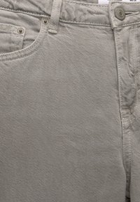 Gros plan sur un jean en denim gris clair montrant la poche avant, la passants de ceinture, le bouton et les coutures texturées du tissu.