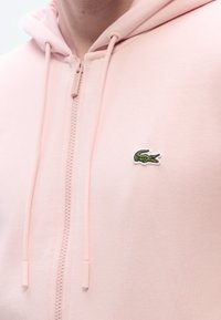 Ροζ φούτερ με φερμουάρ, κατασκευασμένο από μαλακό ύφασμα, με μικρό πράσινο λογότυπο κροκοδείλου στο στήθος και κορδόνια στη κουκούλα.