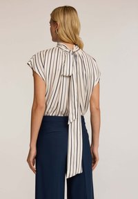 Camicia crema con righe verticali blu navy, maniche corte e un fiocco sul retro del colletto. Indossata con pantaloni blu navy con tasche laterali.