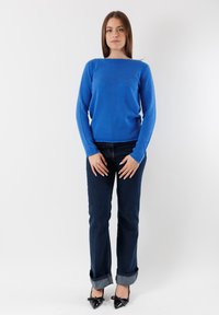 Maglione a maniche lunghe blu in materiale lavorato a maglia, abbinato a jeans blu scuro con orli piegati e scarpe nere a punta con dettagli a fiocco.