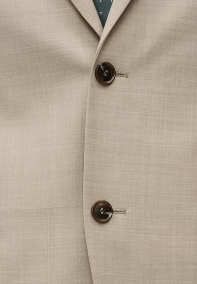 Μπεζ σακάκι από υφασμάτινη υφή, με δύο σκούρα κουμπιά και πράσινη γραβάτα με πουά, ορατή στον γιακά.