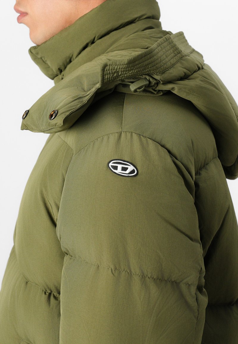 Chaqueta acolchada color verde oliva con un cuello alto, botones a presión, detalles elásticos y un parche de logo en el hombro. Tejido suave y resistente al agua.