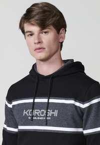 Un tânăr cu părul scurt, de culoare castanie, poartă un hanorac negru și gri cu dungi orizontale și textul "Kuroshi Established in 2001."