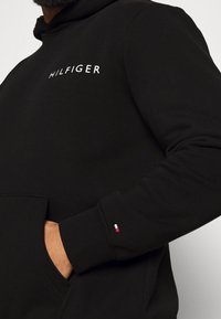 Svart hoodie med vitt "HILFIGER"-logga på bröstet. Har en känguruficka och ribbade ärmslut med en liten röd, vit och blå accent.