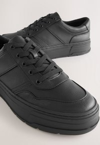 Paire de baskets basses en cuir noir avec semelles épaisses et lacets noirs sur un fond neutre.