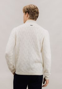 Hvit kabelstrikket genser med høy krage, ribbestrikkede mansjetter og en diskret merkelapp bak. Designet har en teksturert, mønstret konstruksjon.