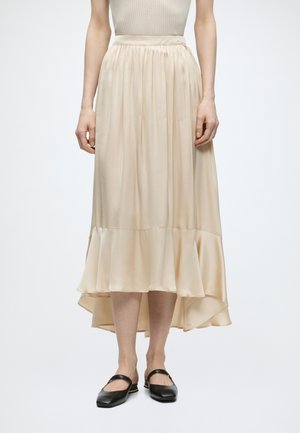 Žena v béžovej sukni so strihaným spodným lemom a čiernymi balerínkami Mary Jane, stojaca na bielom pozadí.