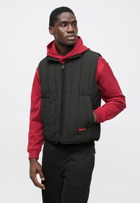 Gilet trapuntato nero con chiusura a zip e logo "HUGO" rosso sul petto, indossato sopra una felpa con cappuccio rossa, abbinato a pantaloni neri.