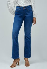 A person stands wearing a white, lacy blouse and blue flared jeans, complemented by brown strappy high heels, against a neutral background.