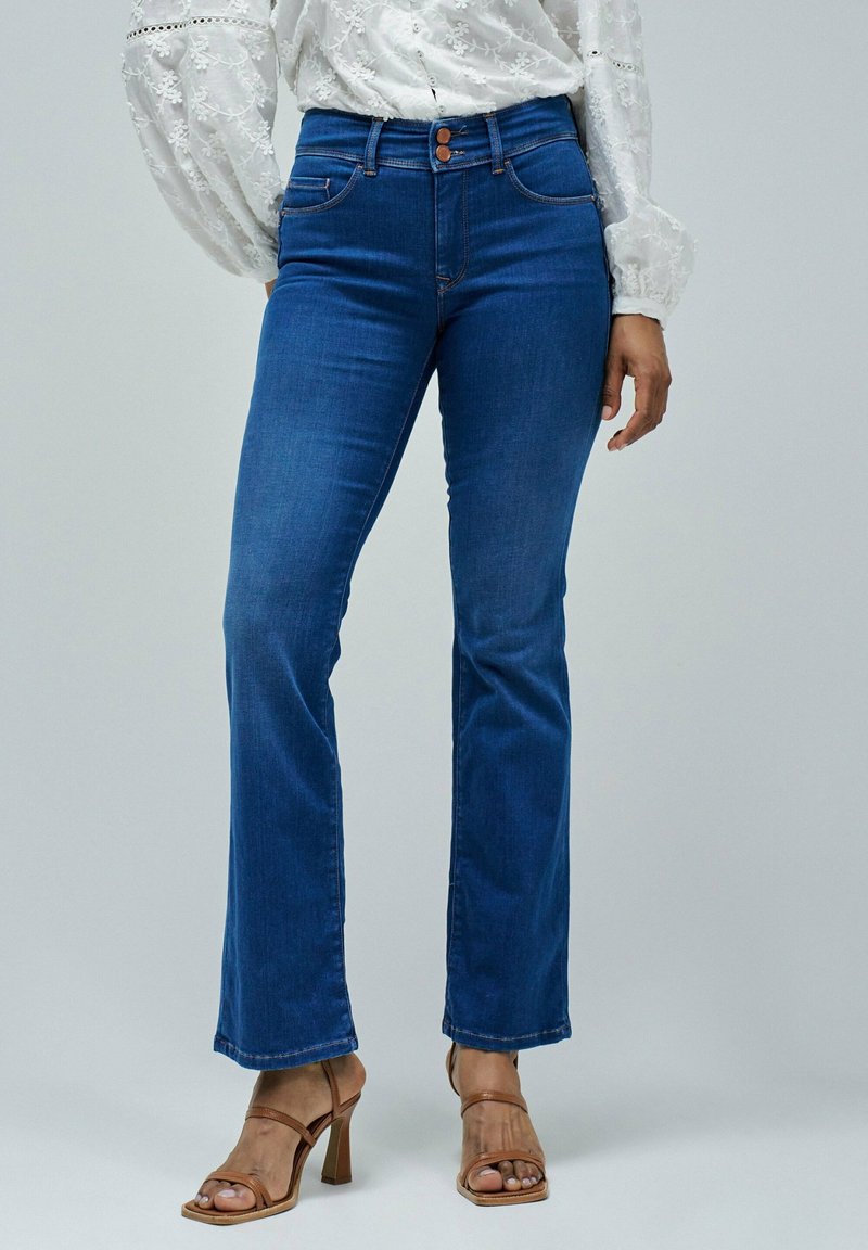 A person stands wearing a white, lacy blouse and blue flared jeans, complemented by brown strappy high heels, against a neutral background.