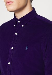 Violetti korduroy-paita, jossa on pitkät hihat, nappikiinnitteinen kaulus ja pieni sininen kirjailtu logo rinnassa; valkoiset napit.