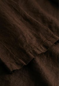 Tissu en lin brun foncé avec une texture et des coutures visibles, présentant des plis et des froissements naturels sous un éclairage doux.