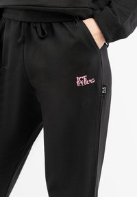 Black sweatpants made of soft fabric, featuring a pink "ICEBERG" logo on the right thigh and a side pocket with a drawstring waist.