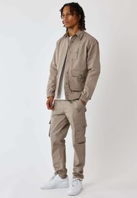 Giacca beige chiaro con bottoni a pressione e più tasche, abbinata a pantaloni cargo coordinati. Sneakers bianche completano l'outfit.