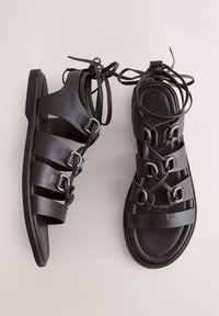 Paire de sandales gladiateur en cuir noir avec plusieurs brides, boucles argentées et lacets enroulés autour de la cheville sur un fond clair.