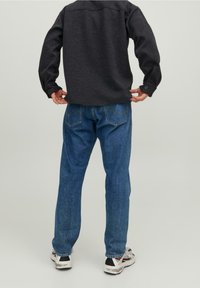 Camicia nera in tessuto intrecciato con maniche lunghe, abbinata a jeans di denim blu; include due tasche posteriori e vestibilità rilassata. Scarpe da ginnastica bianche.