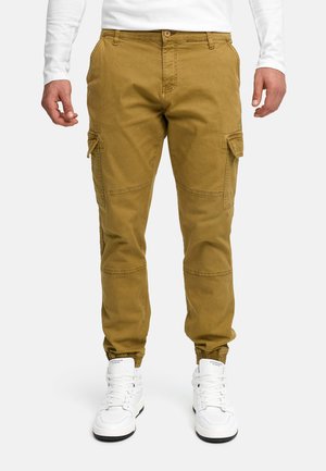 Mann trägt senffarbene Cargo-Hosen mit Seitentaschen und weiße High-Top-Sneaker, steht vor einem weißen Hintergrund.