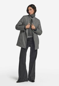 Grå ullkappa med ståkrage, knäppning fram och två sidofickor, bärs över en mörk stickad tröja och utsvängda jeans.