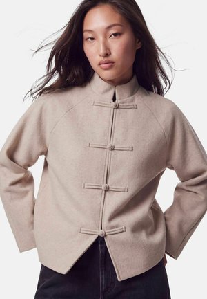 Jeune femme aux longs cheveux foncés portant une veste en laine beige à col montant et quatre boutons noués horizontaux, posant les mains sur les hanches.