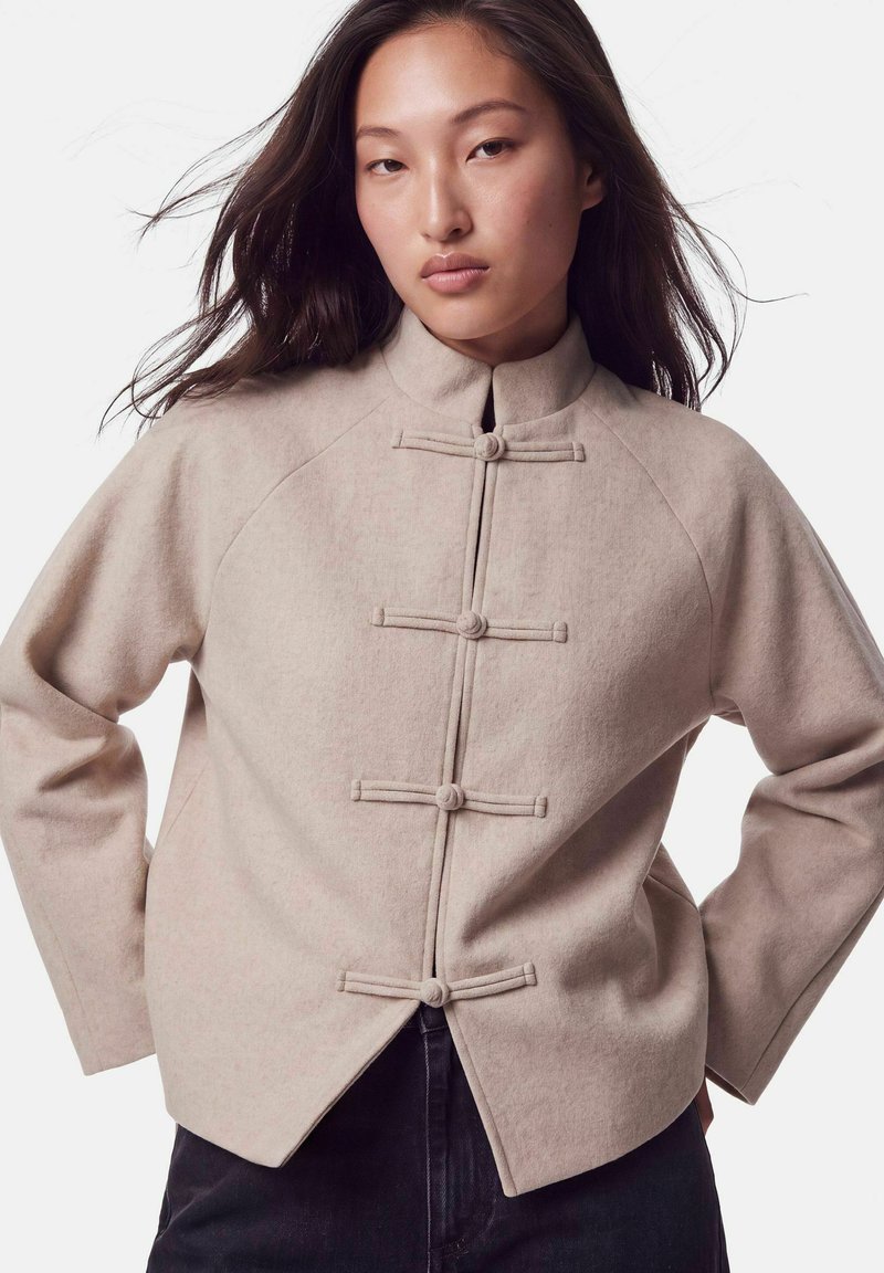 Jeune femme aux longs cheveux foncés portant une veste en laine beige à col montant et quatre boutons noués horizontaux, posant les mains sur les hanches.