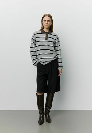 Mujer con polo gris de rayas de manga larga, pantalones cortos anchos negros y botas oscuras hasta la rodilla, de pie frente a una pared clara y lisa.