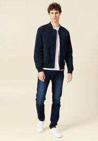 Chaqueta bomber navy, de tejido ligero, con cremallera frontal, puños acanalados, combinada con vaqueros azules y zapatillas blancas, de pie contra un fondo neutro.