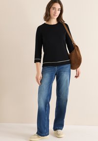 Haut noir à manches longues avec bord blanc, associé à un jean large bleu et un sac à épaule en cuir suédé marron. Des chaussures décontractées complètent le look.