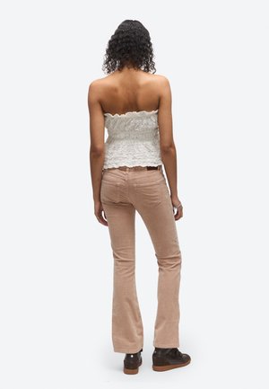 Une femme se tient de dos et présente un haut blanc en dentelle sans bretelles, un pantalon beige évasé et des chaussures foncées sur un fond uni.