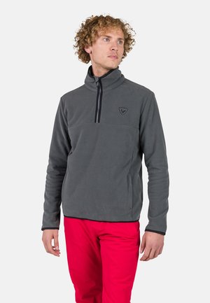 Grijze fleecepullover met hoge kraag en halve rits, voorzien van een zwarte rits en klein logo op de borst, gedragen met rode broek.