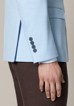 Ręka spoczywająca wzdłuż ciała, ubrana w jasnoniebieską marynarkę z czterema ciemnymi guzikami na mankiecie oraz ciemnobrązowe spodnie.