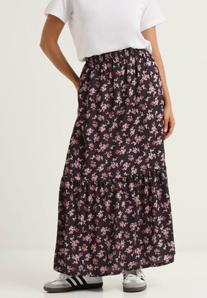 Frau trägt schwarzen Maxirock mit Blumenmuster und geraffter Taille sowie weißen Sneakers, stehend vor einfarbigem Hintergrund.