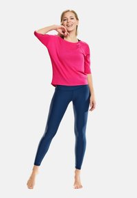Roze top met driekwart mouwen, gecombineerd met glanzende marineblauwe legging. De top heeft een ontspannen pasvorm; de leggings zijn strak en lichaamsvormend.