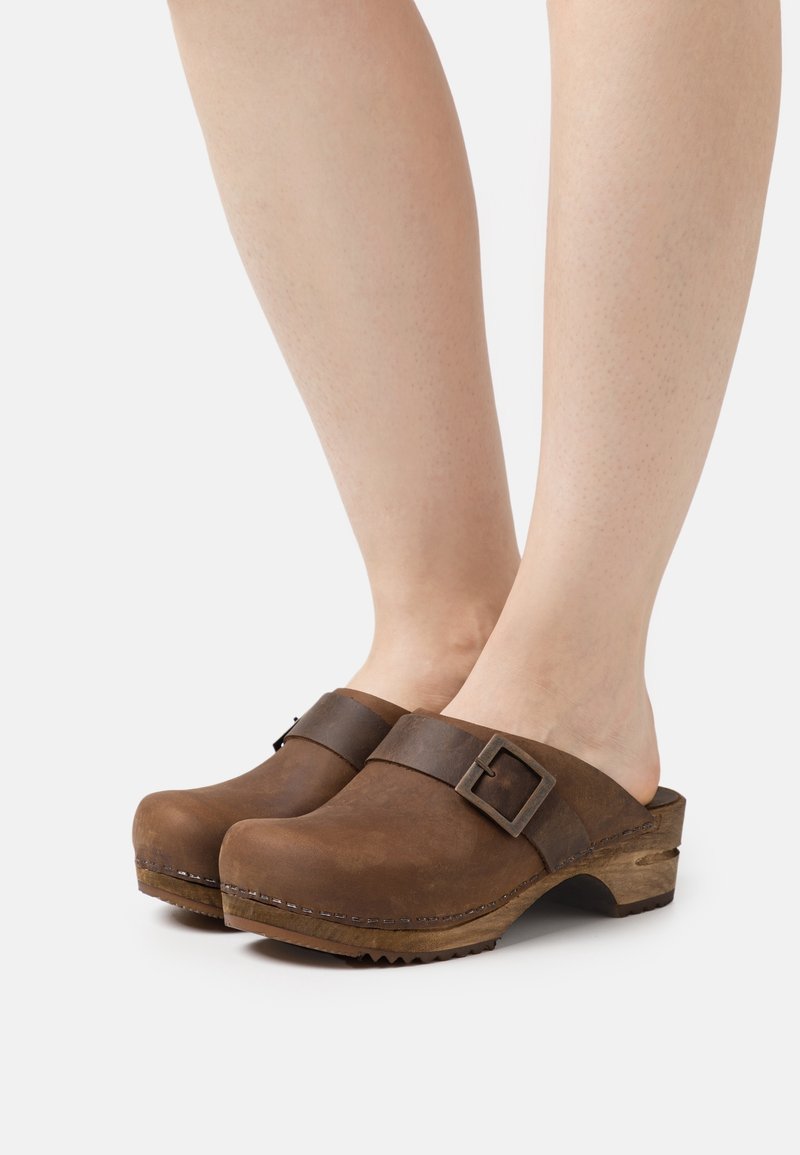 Pieds portant des sabots en cuir marron avec une semelle en bois et une bride réglable à boucle sur fond blanc.