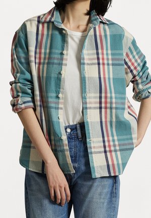 Personne portant une chemise à carreaux bleu sarcelle, crème, rouge et bleu marine aux manches retroussées sur un t-shirt blanc et un jean bleu.