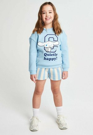 Smiling girl with brown hair wearing a blue sweatshirt with cartoon dog and "Quietly happy!" text, striped shorts, white socks, and sneakers.