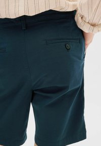 Pantaloni corti blu scuro realizzati in tessuto misto cotone, dotati di una tasca posteriore con chiusura a bottone e una texture liscia. Taglio sartoriale.