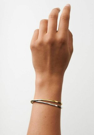 Mano destra con dita leggermente piegate, indossa due sottili braccialetti ondulati, uno oro e uno argento, su uno sfondo bianco uniforme.