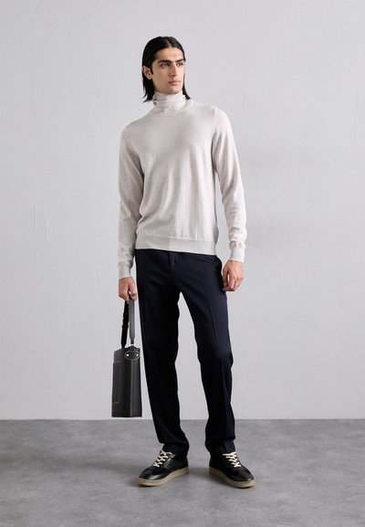 Maglione a collo alto grigio chiaro, pantaloni blu navy, sneakers nere, tiene una borsa grigia scura. Texture liscia, design aderente e sottile contrasto di colore.