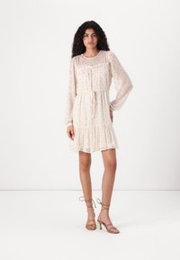 Robe beige clair avec de longues manches transparentes, motif à pois, encolure froncée, taille élastique et jupe à volants. Associée à des sandales à talons hauts avec des lanières.