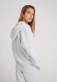 Lichtgrijze hoodie van zacht fabric, met een grote capuchon, losse pasvorm en ribgebreide boorden. Model is zijwaarts afgebeeld en draagt bijpassende broek.