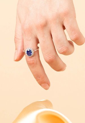 Silver ring with an oval blue gemstone surrounded by small clear stones, worn on a hand. Background is a soft beige color.