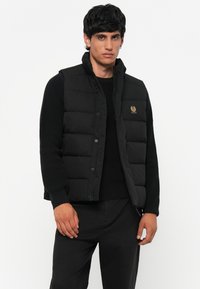 Black puffer vest with a high collar and snap buttons, featuring a small gold emblem. Worn over a black sweater, with a textured finish.