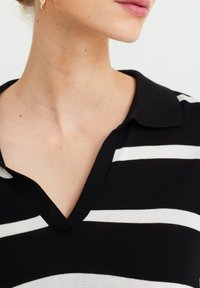 Černobílý pruhovaný polo s designem do V, vyrobený z jemného materiálu a s límcem. Viditelné detaily šití kolem výstřihu.