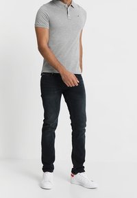 Grijze polo met korte mouwen en drie-knopssluiting, gecombineerd met een donkere spijkerbroek en witte sneakers met rode accenten. Effen achtergrond.
