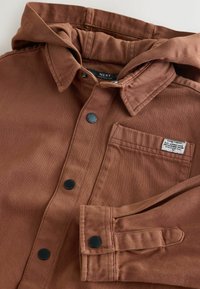 Veste marron à boutons avec capuche. Elle comporte des boutons pression noirs et une poche poitrine. Tissu texturé avec des détails de couture visibles.